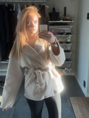 Beige kappa missäy - Stilren beige kappa med bred krage och knytskärp i midjan. Modellen har en avslappnad passform och vida ärmar, perfekt för lager på lager. Tillverkad i ett mjukt tyg som ger en lyxig känsla. Passar till både jeans och klänning för en trendig look.