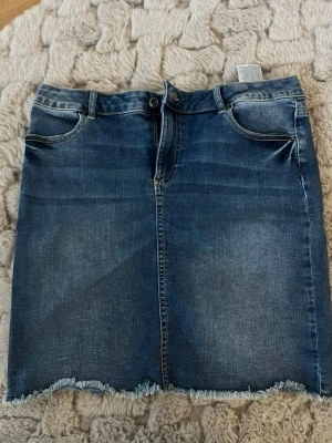 Blå jeanskjol med rå kant - Snygg blå jeanskjol i klassisk modell med fem fickor och rå, fransig kant nedtill. Kjolen har normal passform och stängs med knapp och dragkedja framtill. Perfekt till vardagslooken och enkel att matcha med olika toppar.