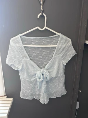 Ljusblå mesh blus med knyt framtill - Söt ljusblå blus med korta ärmar och knyt framtill. Blusen har ett diskret blommigt mönster i tyget och är lätt transparent förutom vid bysten