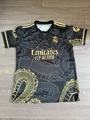 Real Madrid svart matchtröja drake - Snygg svart Real Madrid matchtröja med guldiga detaljer och stort drakmönster. Tryck med 'Emirates Fly Better' på bröstet och 'VINI JR. 7' på ryggen. Korta ärmar, rund hals och LA LIGA-märke på ärmen. Perfekt för fotbollsfans som vill sticka ut.