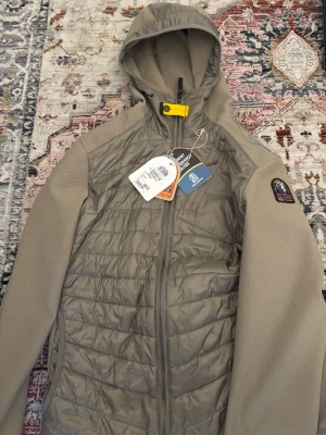 Beige vindjacka från Parajumpers - Snygg beige/grön vindjacka från Parajumpers med huva och dragkedja framtill. Jackan har quiltad front, släta ärmar och en gul detalj vid halsen. Märkespatch på ärmen och flera praktiska fickor. Materialet är en mix av syntet och mesh för extra komfort. 100% äkta