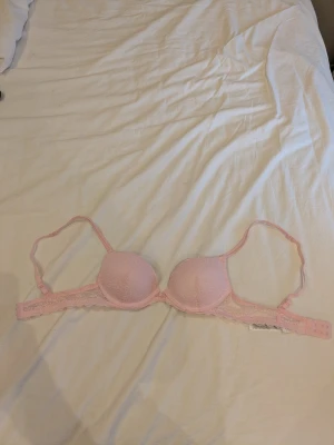 Ljusrosa spets-bh med vaddering - Söt ljusrosa bh med vadderade kupor och spetsdetaljer. Justerbara axelband och hakknäppning bak. Den är i storlek 70A.