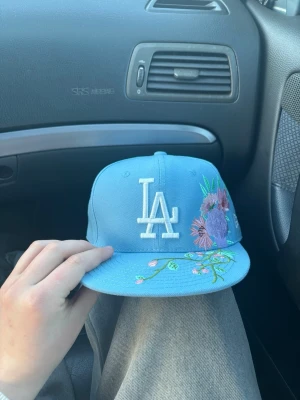 Blå LA keps med blommor från New Era - Säljer en ljusblå snapback-keps från New Era med LA-logga framtill i vitt och broderade blommor i lila, rosa och grönt på sidan och skärmen. Klassisk rak skärm och justerbar baksida. Perfekt för dig som vill sticka ut med en unik accessoar.