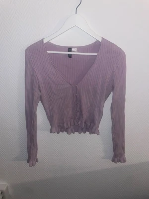 Ljuslila ribbad kofta från H&M Divided - Säljer en ljuslila ribbad kofta från H&M Divided. Koftan har v-ringning, långa ärmar och knappar framtill. Den är croppad med volangkant nedtill och vid ärmsluten, vilket ger en söt och trendig look. Perfekt att matcha med jeans eller kjol.