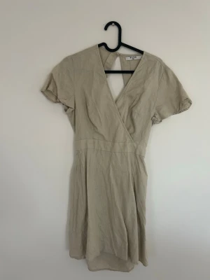 Beige klänning från NA-KD - Snygg beige klönning från NA-KD i luftigt linnematerial. Klänningen har korta ärmar, v-ringning och en avslappnad passform som ger en stilren och enkel look. Perfekt för varma dagar och enkel att styla med accessoarer.