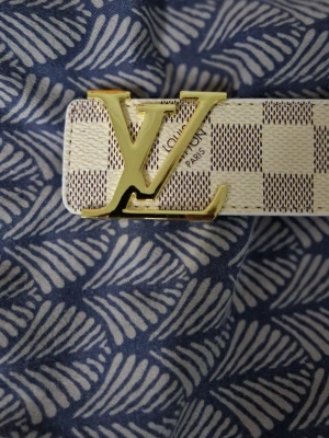 L v Damier-bälte beige - Snyggt L V-bälte i beige och vitt Damier-mönster med stor guldfärgad L V-logga som spänne. Bältet är smalt och har en exklusiv känsla, perfekt för att lyfta vilken outfit som helst. Materialet är canvas med detaljer i metall.