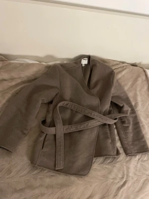 Brun  kappa i ullmix med knytskärp - Stilren brun kappa i ullmix med avslappnad passform och knytskärp i midjan. Kappan har en öppen front utan knappar och raka ärmar. Perfekt för lager-på-lager och enkel att styla till olika outfits.