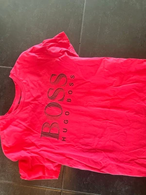 Hugo Boss t-shirt - Snygg neonrosa t-shirt från Hugo Boss med stort svart logotryck på bröstet. Klassisk rund halsringning och korta ärmar. Perfekt för dig som vill sticka ut och gillar streetwear vibes.