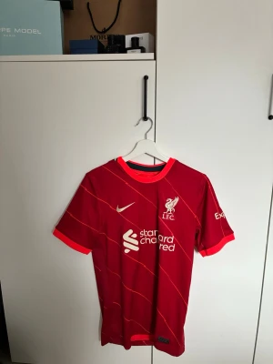 Liverpool matchtröja röd Nike - Snygg röd Liverpool FC matchtröja från Nike med vita och orange detaljer. Tröjan har klubbmärke och sponsortryck på bröstet samt korta ärmar. Tillverkad i lätt och ventilerande material, perfekt för fotboll eller träning.