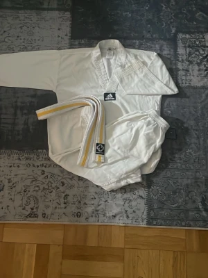 Vit taekwondodräkt från Adidas tillsammans med ett bälte - Säljer en klassisk vit taekwondodräkt från Adidas med långärmad överdel, byxor med resår i midjan och ett vitt bälte med gula ränder. Dräkten har V-ringad krage och är tillverkad i ett lätt och slitstarkt material, perfekt för träning och tävling.