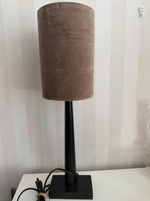 Bordslampa med brun lampskärm Havsö - Snygg bordslampa med cylinderformad brun lampskärm i tyg och svart metallfot. Lampan har en fyrkantig bas och svart sladd med strömbrytare. Passar perfekt på skrivbord eller nattduksbord för en stilren look. Skärm tryckt dock inget som syns på utsidan, höjd 45 cm.