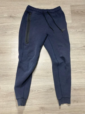 Mörkblå Nike tech byxor  - Snygga mörkblå mjukisbyxor från Nike med smal passform, resår i midjan och svarta dragsnören. Byxorna har en dragkedjeförsedd ficka på sidan och ribbade muddar vid bensluten. Perfekta för chill eller träning.