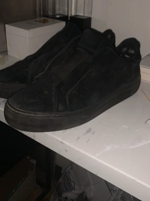 Svarta slip-on sneakers i mocka - Säljer ett par helsvarta slip-on sneakers i mocka med minimalistisk design. Skorna har en låg siluett och saknar synliga snören, vilket ger en clean och stilren look. Perfekta för dig som gillar enkelhet och streetwear-vibe.