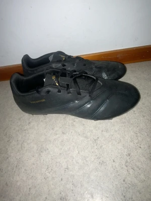 Adidas Predator svarta fotbollsskor - Säljer ett par svarta Adidas Predator fotbollsskor med klassisk design och snörning. Skorna har guldiga detaljer och är tillverkade i syntetmaterial. Perfekta för dig som vill ha stilrena och funktionella skor på planen.