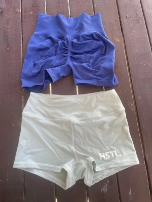Två par träningstights shorts - Säljer två par träningstights shorts, ett par i blått med hög midja och ett par i ljusgrått med HSTL.-logga. Båda är stretchiga och perfekta för gym eller löpning. Snygg passform och bekvämt material som andas. Båda för 350kr. Endast swish❤️🫶