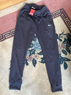 Mörkgrå/Svart Nike Miller byxor - Snygga mörkgrå träningsbyxor från Nike med smal passform och resår i midjan. Byxorna har dragsko, sidofickor och ribbade muddar vid bensluten. Tillverkade i ett mjukt och stretchigt material som passar perfekt till träning eller chill.