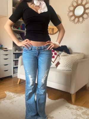 Ljusblå bootcut jeans med slitningar - Säljer ett par ljusblå bootcut jeans med slitningar framtill och broderade detaljer på bakfickorna. Jeansen har låg midja och klassisk femficksdesign. Perfekta för dig som gillar en avslappnad men trendig look.