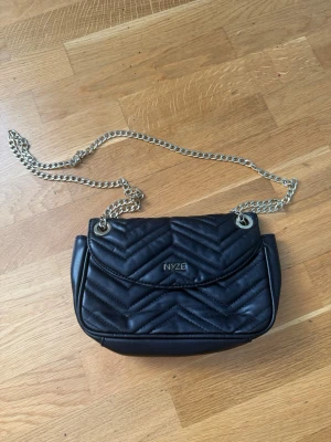 Svart quiltad handväska från NYZE - Snygg svart quiltad handväska från NYZE med silverfärgad kedjerem och elegant mönster på framsidan. Väskan har lock med magnetknäppning och detaljer i metall. Perfekt för dig som vill ha en trendig accessoar med plats för det viktigaste.