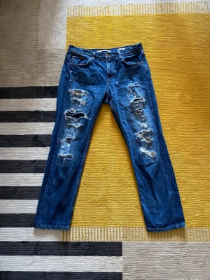 Premium Salvadge jeans - Säljer ett par blå jeans med rejält slitna detaljer och stora hål på framsidan av benen. Jeansen har klassisk femficksmodell och raka ben. Perfekt för dig som gillar en edgy och trendig stil. Materialet är denim i bomull.