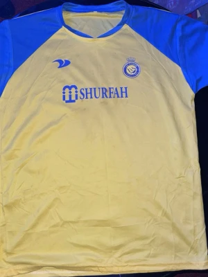 Al Nassr Ronaldo matchtröja gul/blå - Snygg gul matchtröja från Al Nassr med blå detaljer och tryck. Framsidan har klubbmärke och sponsorlogga, baksidan har namnet 'Ronaldo' och nummer 7 i blått. Tillverkad i lätt och ventilerande material, perfekt för fotboll eller träning.