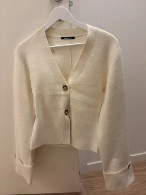 Kofta från Gina Tricot - Säljer en krämvit kofta från Gina Tricot med v-ringning och två stora knappar framtill. Koftan har långa ärmar med dekorativa knappar vid ärmsluten och en något boxig, kortare passform. Perfekt att styla med jeans eller kjol