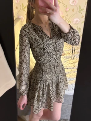 Mönstrad kort klänning - Säljer en snygg kort klänning med leopard aktigt mönster. Klänningen har långärmad, transparenta ärmar, markerad midja med smock och volangig kjol. V-ringad framtill med snörning och tillverkad i ett lätt, luftigt material som ger en skön sommar vibe. Tyget har små skimmriga detaljer. Perfekt nu till sommaren. Säljer då den tyvärr blivit lite för liten.