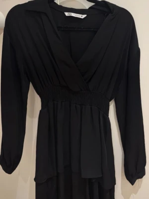 Svart omlottklänning från Zara - Säljer en svart omlottklänning från Zara med v-ringning och långa ärmar. Klänningen har smock i midjan för en snygg passform och ett lätt, flytande tyg som ger en elegant look. Perfekt för dig som gillar stilrena plagg med fina detaljer.