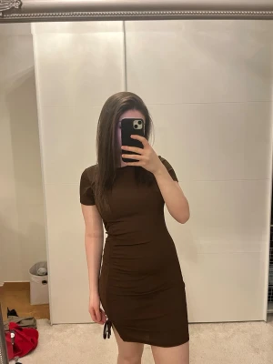 Brun kort klänning med snörning - Säljer en brun kortärmad klänning från H&M med snörningsdetalj på sidan. Klänningen är figurnära och har en lätt rynkad effekt längs ena sidan. Perfekt för dig som gillar en enkel men trendig look.