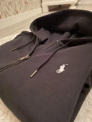Svart hoodie från Ralph Lauren - Säljer en svart hoodie från Ralph Lauren med dragkedja och klassisk vit logga på bröstet. Hoodien har justerbar huva med snören och känguruficka framtill. Tillverkad i mjukt bomullsmaterial, perfekt för en chill och stilren look.