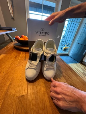 Valentino Garavani vita sneakers - Snygga vita sneakers från Valentino Garavani med bred svart detalj på sidan och vita snören. Skorna har en chunky sula och nitar på hälen för extra edge. Tillverkade i läder och har en klassisk låg profil som passar till det mesta.