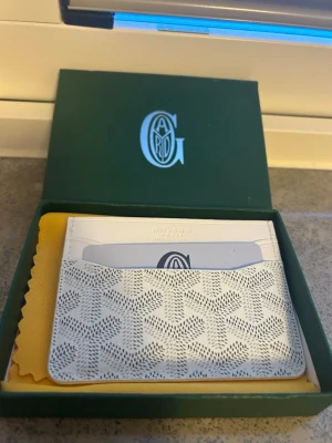 Vit korthållare - Snyggt vitt kortfodral med det klassiska mönstret och loggan tryckt på framsidan. Tillverkad i exklusivt läder och har flera fack för kort. Kommer i original grön ask och med gul putsduk. Perfekt för dig som vill ha stilren och lyxig accessoar.