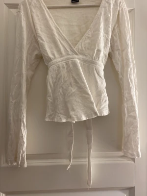 Vit omlottopp med långa ärmar - Säljer en vit omlottopp med långa ärmar och knytband i midjan. Toppen har djup v-ringning och är gjord i ett lätt, strukturerat tyg som ger en avslappnad men ändå stilren look. Perfekt att styla med jeans eller kjol för en trendig outfit.💓
