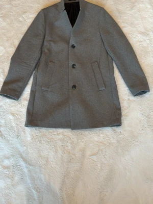 Grå klassisk ullrock med knappar - Stilren grå ullrock med tre knappar framtill och två snedställda fickor. Rak passform och enkel design som ger en tidlös look. Perfekt för kyligare dagar och funkar till många olika outfits.