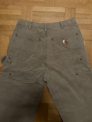 Grå cargobyxor från Carhartt - Säljer ett par grå cargobyxor från Carhartt med flera praktiska fickor, hammarögla på sidan och förstärkta sömmar. Byxorna har raka ben och är tillverkade i slitstark bomullscanvas. Perfekta för en avslappnad och street-inspirerad stil.
