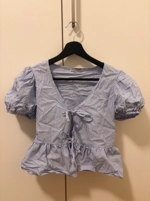 Blårandig blus med puffärm och knyt - Söt blus med blå och vita ränder, puffärmar och v-ringning. Framtill finns dekorativa knytband och volang nedtill. Perfekt för dig som gillar en romantisk och trendig stil.