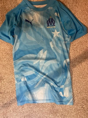 Ljusblå Marseille matchtröja Puma - Säljer en ljusblå matchtröja från Olympique de Marseille med abstrakt mönster och klubbens logga på bröstet. Tröjan har korta ärmar, rund hals och är tillverkad i ett lätt, andningsbart material som passar perfekt till träning eller match.