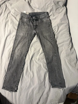 Levis 501 grå - Säljer ett par grå jeans från Levis med rak passform och klassisk femficksdesign. Jeansen har bälteshällor, knappgylf och är tillverkade i ett slitstarkt denimtyg. Perfekta för en avslappnad och trendig look.