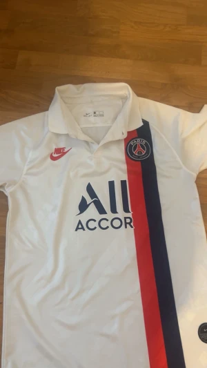 PSG vit matchtröja från Nike - Snygg vit PSG matchtröja från Nike med röd och marinblå rand på framsidan. Tröjan har korta ärmar, krage och Paris Saint-Germain-logga på bröstet. Tillverkad i ett lätt och ventilerande material, perfekt för fotboll eller träning.