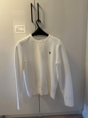 Vit sweatshirt från Polo Ralph Lauren - Klassisk vit sweatshirt från Polo Ralph Lauren med rund halsringning och den ikoniska lilla broderade loggan på bröstet. Tillverkad i mjuk bomullsblandning och har ribbade muddar vid ärmslut och nederkant. Perfekt för en clean och stilren look.