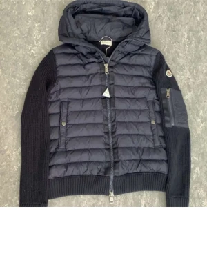 Mörkblå cardigan moncler  - Snygg mörkblå cardigan från Moncler med stickade ärmar och huva. Jackan har quiltad front, dragkedja, fickor med tryckknappar och en extra ficka på ärmen. Klassisk Moncler-logga på armen och ribbade muddar för extra komfort.
