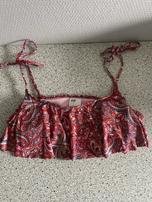 Mönstrad bikiniöverdel från H&M - Säljer en färgglad bikiniöverdel från H&M med paisleyinspirerat mönster i rött, rosa och blått. Modellen har volangdetaljer framtill och tunna axelband med knyt. 