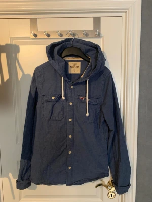 Hollister overshirt  - Blå overshirt  från Hollister med huva och vita snören. Skjortan har två bröstfickor med knappar och Hollister-logga broderad på ena fickan. Långärmad och tillverkad i bomull, perfekt för en avslappnad stil.