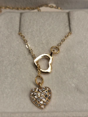 Guldigt halsband med hjärthängen - Snyggt guldigt halsband med två hjärtformade hängen, varav det ena är öppet och det andra är täckt med glittrande stenar. Kedjan är tunn och elegant, perfekt för att ge en romantisk touch till din outfit. Kedja 48 cm + 5 cm förlängningskedja. Hängena är ca 1 cm. 