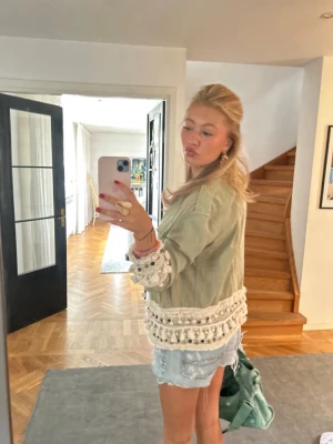 Grön overshirt med virkade detaljer - Snygg grön overshirt med virkade spetsdetaljer och tofsar vid ärmslut och nederkant. Jackan har knappar framtill och en bröstficka. Perfekt för dig som vill ha en unik och trendig look.