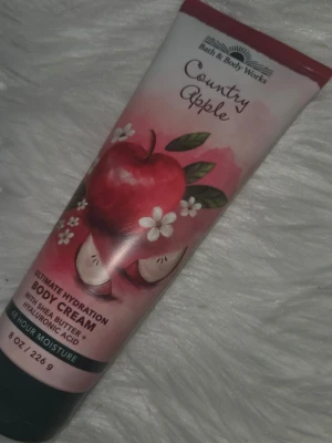 Bath & Body Works Country Apple Body Cream - Fuktgivande body cream med doft av äpple, sheasmör och hyaluronsyra. Tuben är röd och vit med äppelmönster och innehåller 226 g. Ger 48 timmars återfuktning och lämnar huden mjuk och len.
