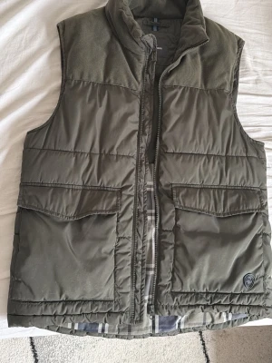 Olivgrön dunväst från Dressmann - Snygg olivgrön dunväst från Dressmann med hög krage och fyra stora fickor framtill. Västen har quiltad design och dragkedja. Materialet är glansigt och känns slitstarkt, perfekt för lager-på-lager-stil. Märkeslogga på ena fickan ger en extra detalj.