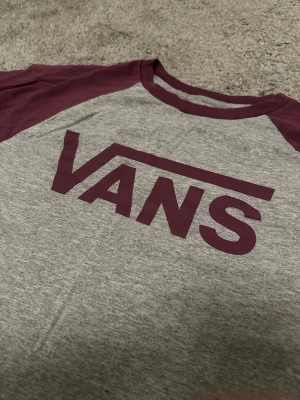 Grå och vinröd Vans t-shirt - Säljer en grå t-shirt från Vans med vinröda trekvartsärmar och matchande tryck på bröstet. Klassisk baseballmodell med rund halsringning och avslappnad passform. Perfekt för dig som gillar streetwear och enkel stil.