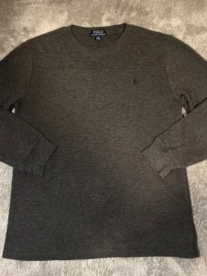 Mörkgrå långärmad tröja Polo Ralph Lauren - Mörkgrå långärmad tröja från Polo Ralph Lauren med rund halsringning och diskret broderad logga på bröstet. Tröjan är ribbstickad vid ärmsluten och har en klassisk, stilren design som passar till det mesta. Tillverkad i mjukt material för extra komfort. Skick 10/10 Nypris - 1499kr