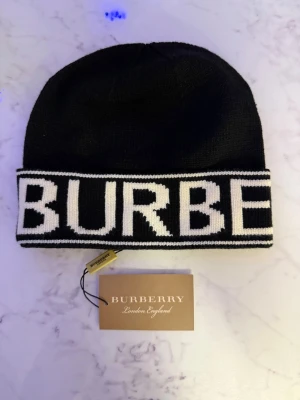 Svart mössa från Burberry - Snygg svart mössa från Burberry med bred vit kant där BURBERRY är stickat i stora bokstäver. Klassisk beanie-modell i mjukt stickat material, perfekt för att hålla värmen och ge en trendig look.