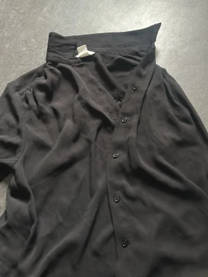 Svart skjorta med knappar - Stilren svart skjorta med klassisk krage och knäppning framtill. Skjortan har lång ärm och är tillverkad i ett mjukt, tunt material som ger ett snyggt fall. Perfekt att styla till olika looks.